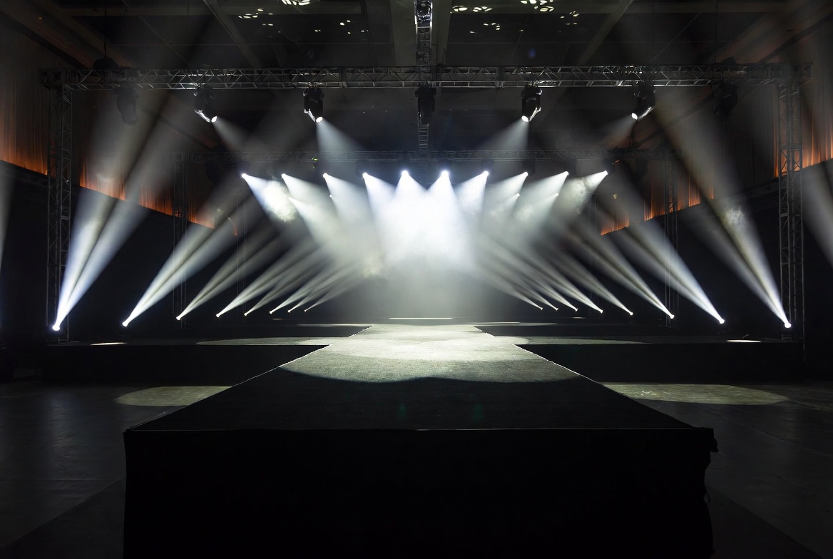 Dramatic empty runway show set with overhead spotlights, polished catwalk surface, and audience seating prepared for fashion event