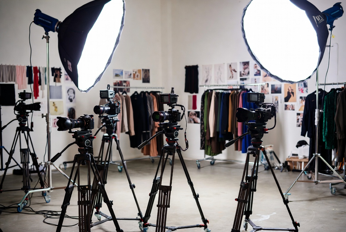 Active fashion production set with professional cinema cameras, lighting fixtures, wardrobe styling area, and crew working on a campaign shoot