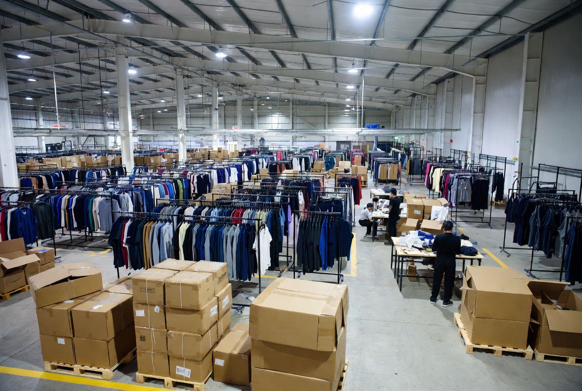 Garment factory warehouse with finished clothing inventory hanging on rolling racks adjacent to packaging stations