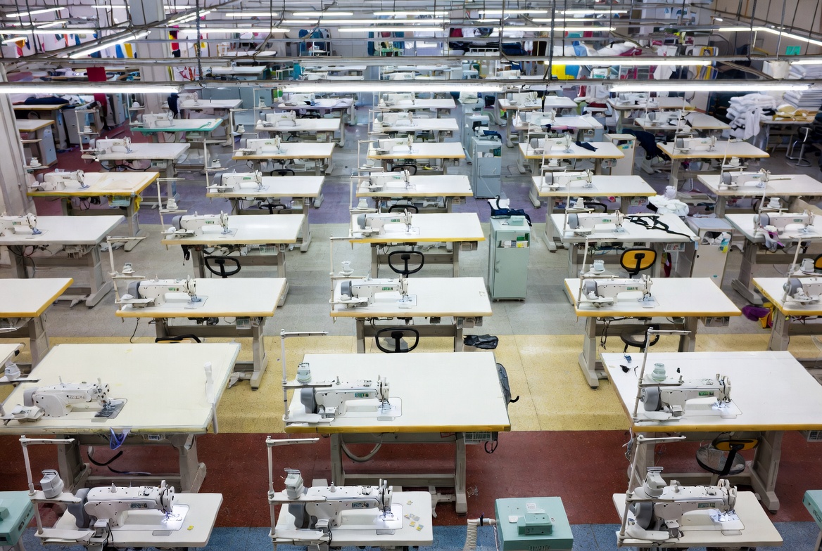Industrial garment contractor production floor with rows of industrial sewing machines, fabric stations, overlock and bartack equipment, and a working production crew